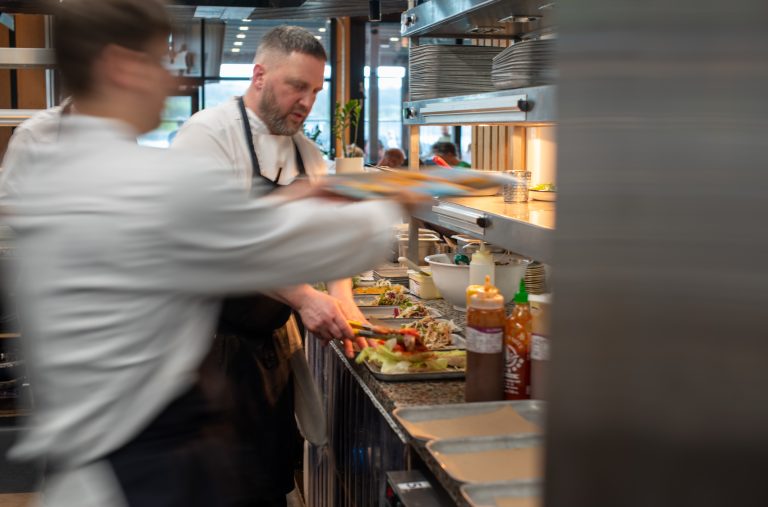 Chefs in a bustling kitchen, plating dishes with focus and precision, reminiscent of the dynamic energy found at food wholesale markets. The blurred motion of one chef adds a sense of urgency and activity.